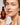 Eine Frau mit heller Haut und lockigem Haar lächelt sanft in die Kamera und hält eine braune Flasche Farm Loves Face Ceramide + Bio-Chicoree Barriere Hydro Creme hoch, eine Feuchtigkeitscreme zur Stärkung der Hautbarriere.